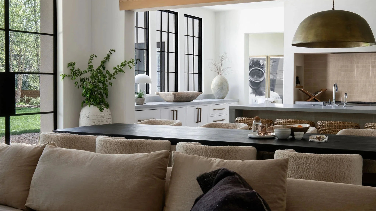 A modern living room interior featuring a beige bouclé sofa in the foreground, a dark wood dining table, and an open kitchen area in the background with black-framed windows and a brass pendant light.