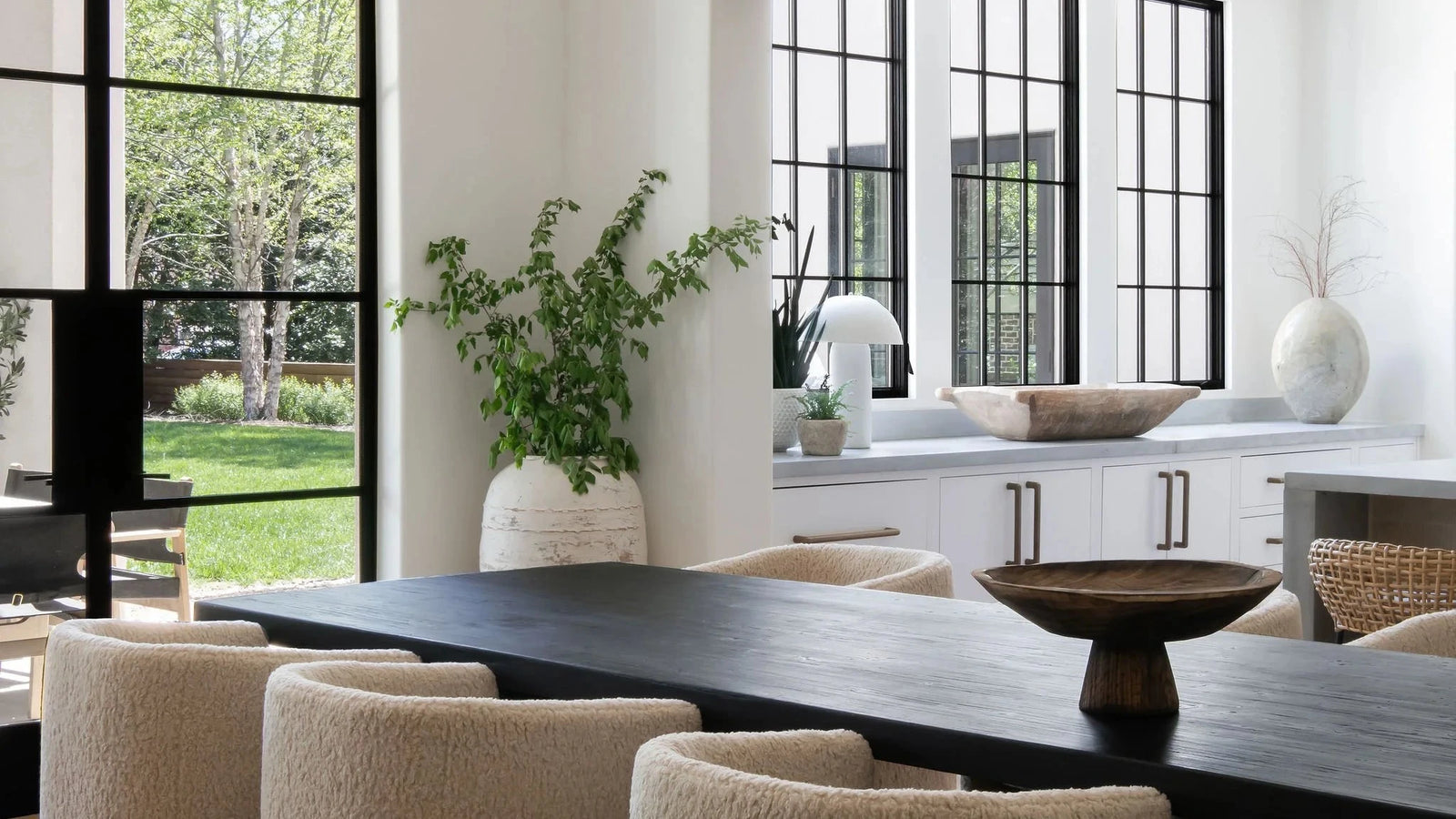 A modern dining room interior with a dark wood table and textured beige upholstered chairs, set in a bright home with large black-framed windows and a visible outdoor green lawn.