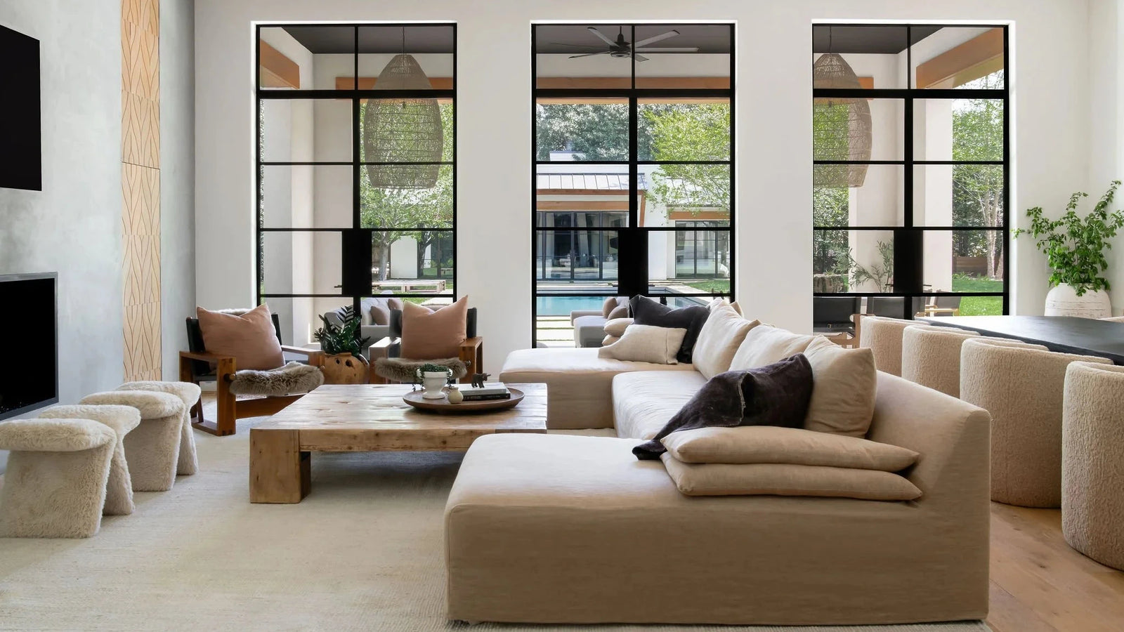 A modern beige sectional sofa with a black cat resting on it, set in a bright contemporary living room with large black-framed windows, a wooden coffee table, and textured beige stools.