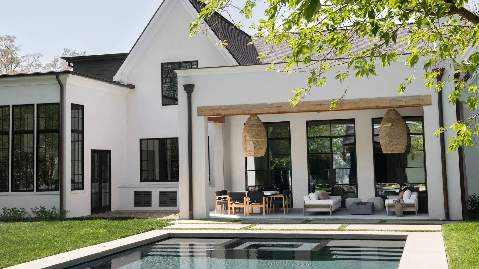 Modern white house exterior with a rectangular swimming pool in the foreground, large black-framed windows, outdoor seating under a covered patio, and green lawn with tree branches overhead.