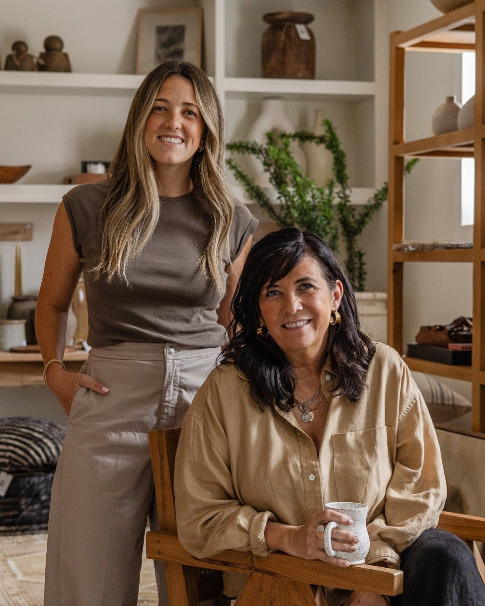 Two women posing in a warm, rustic pottery studio filled with wooden shelves and ceramic vessels. The woman standing wears a taupe sleeveless top and light beige pants, while the seated woman wears a beige button-down shirt and holds a white mug.