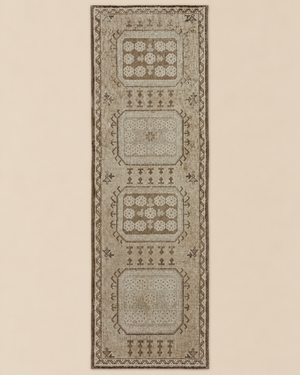 A long rectangular area rug with a distressed traditional pattern in beige, taupe, and brown tones, shown flat against a solid light beige background.
