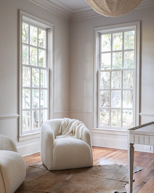 A styled interior room corner with two tall white-framed windows, a cream boucle armchair with a draped throw, part of a matching cream chair at left, and a brown cowhide rug on hardwood floors.