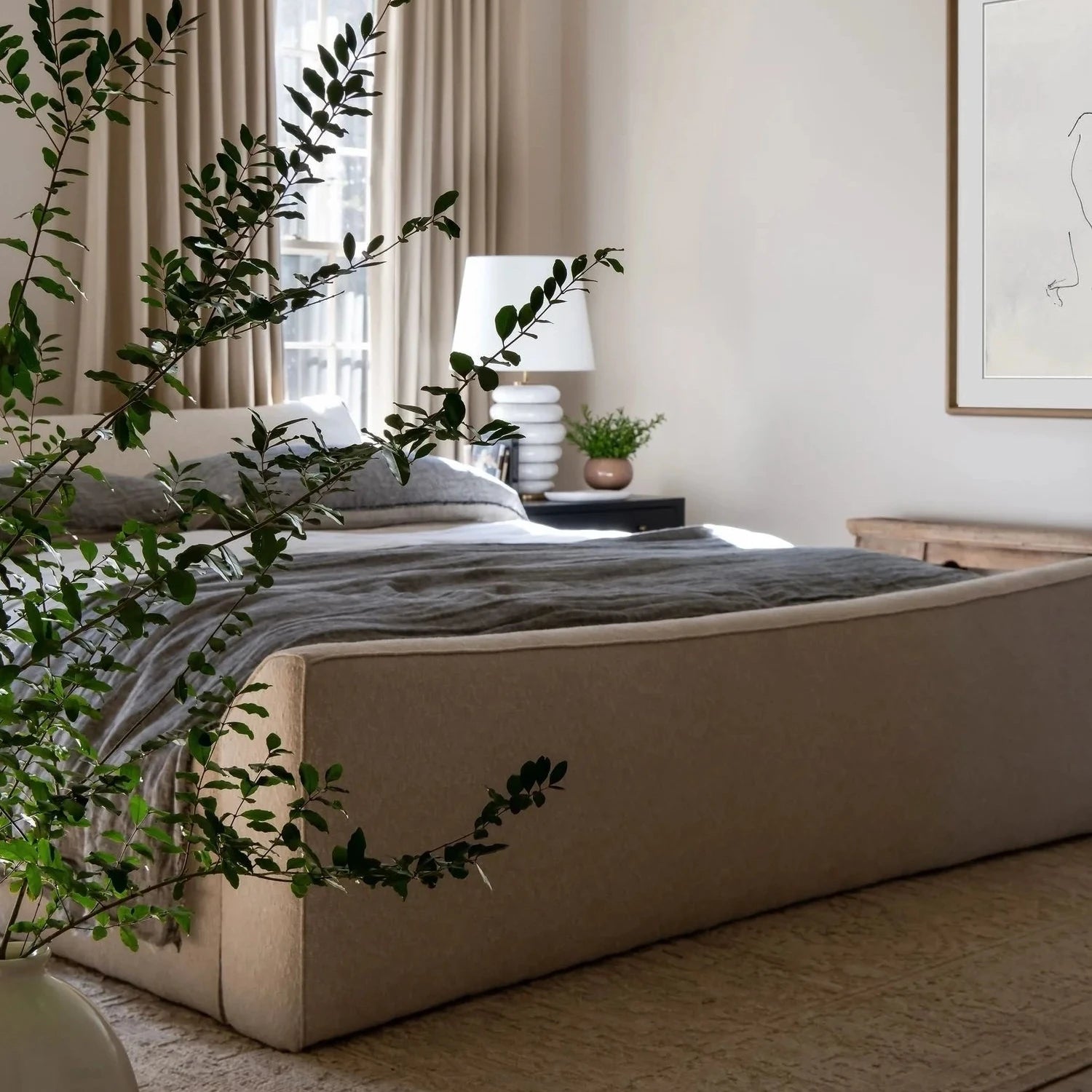 A modern bedroom featuring a low-profile upholstered platform bed in a warm taupe-beige fabric, styled with neutral beige curtains, a matching rug, and a dark gray textured throw blanket. A large green plant in the foreground and a hanging dome-shaped pendant light frame the scene.