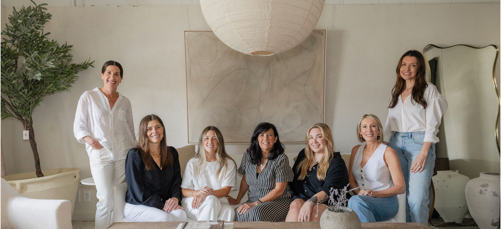 Seven women posing together in a bright, modern living room with a large wooden table, white paper lantern, and neutral-toned wall art.