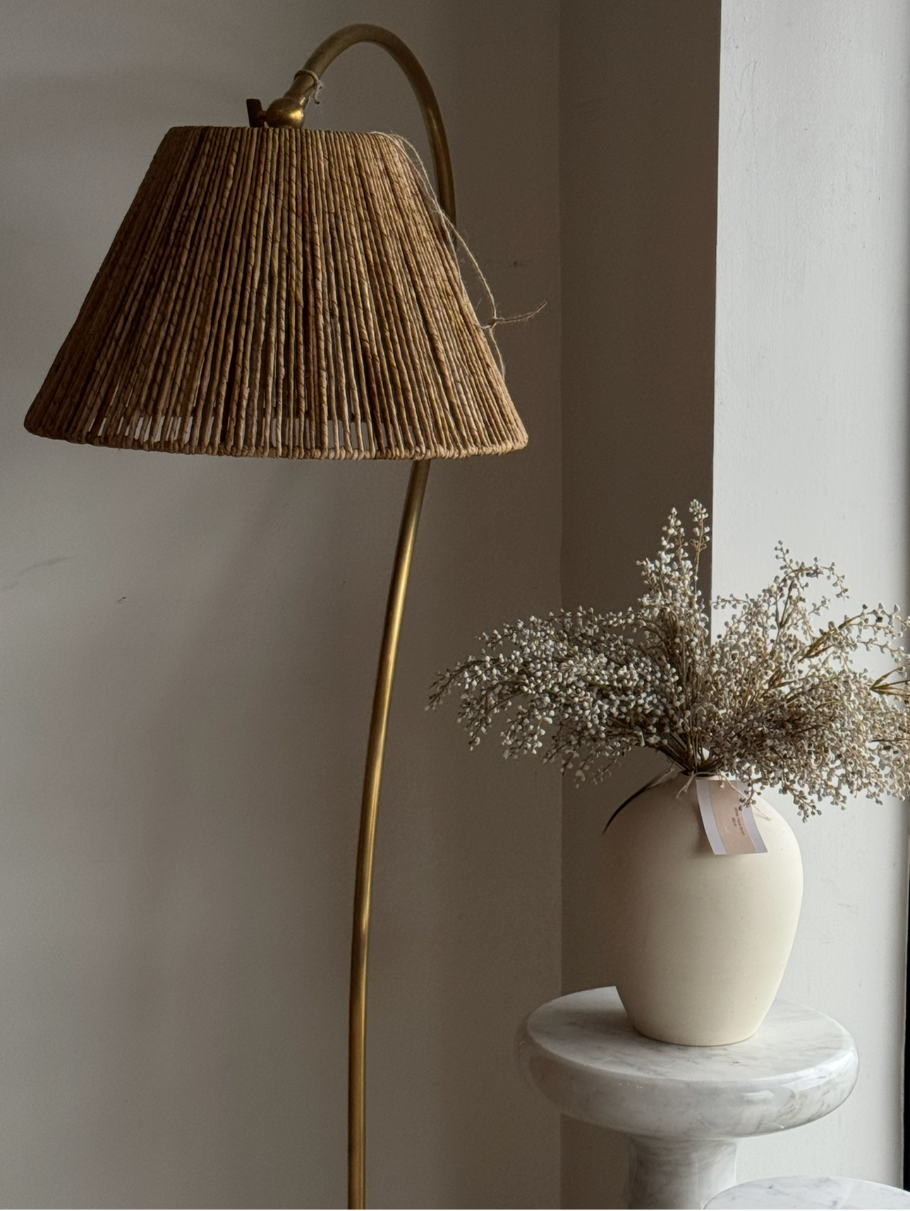 A brass floor lamp with a pleated natural jute shade stands beside a cream ceramic vase filled with dried baby's breath on a white marble pedestal against a neutral wall.