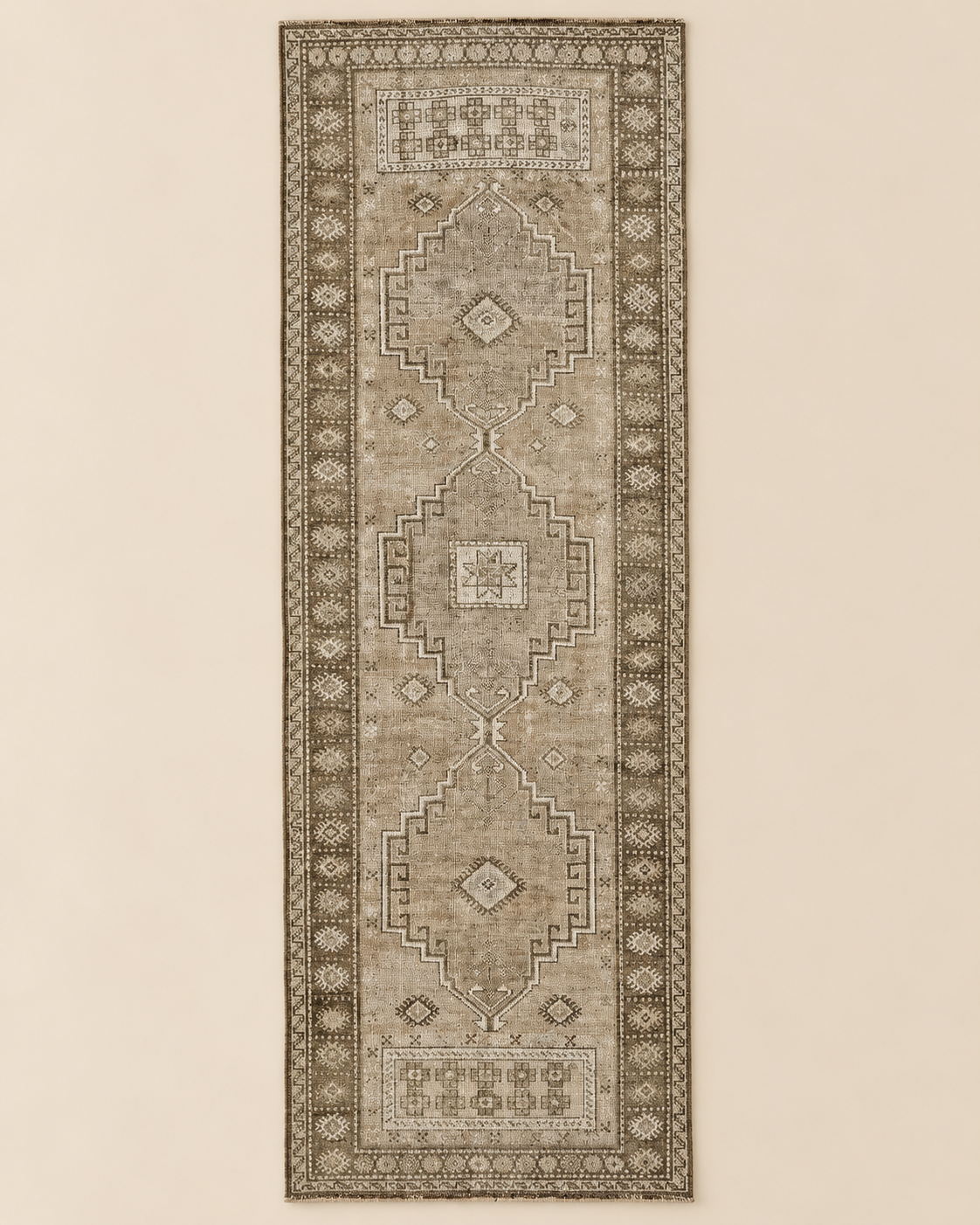 A long rectangular area rug with a distressed traditional medallion pattern in muted beige, taupe, and brown tones, shown flat against a light beige background.