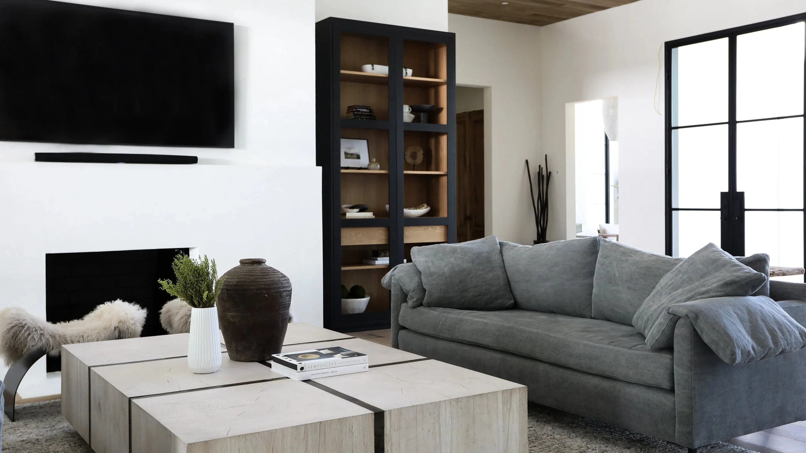 A modern living room featuring a gray fabric sofa, a light wood coffee table, a black wall-mounted TV, and a tall black shelving unit with wood interior.