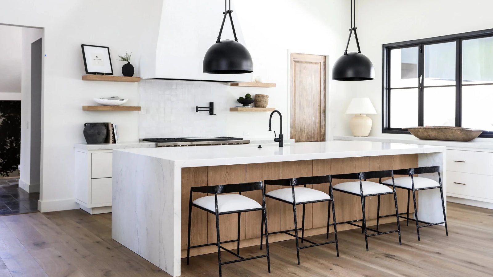 A modern kitchen island with a white marble-look top and base, light wood cabinetry, and five white bar stools with black metal frames. Two black pendant lights hang above the island, set in a bright kitchen with white walls, wood floors, and a large window.