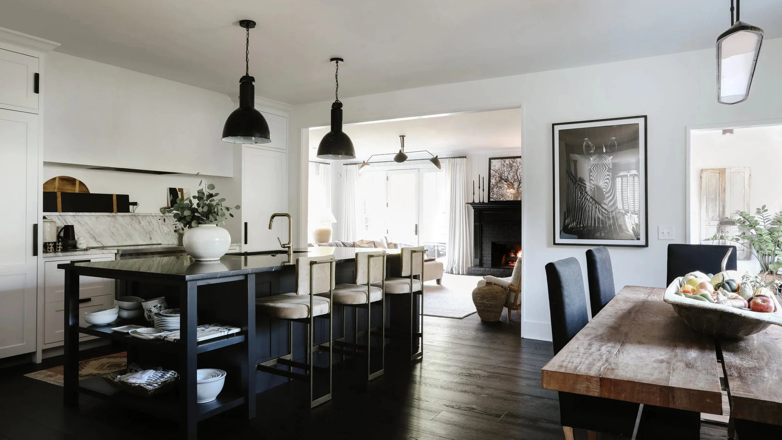 A modern open-plan kitchen and dining area with a dark wood island, black pendant lights, a rustic wooden dining table, and dark chairs, set in a bright contemporary home.