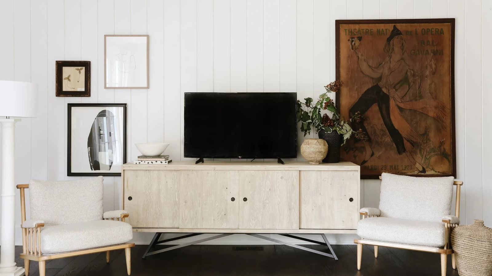 A modern light wood media console in a bright living room setting, styled with a black flat-screen TV, a potted plant, framed wall art, and two white armchairs.