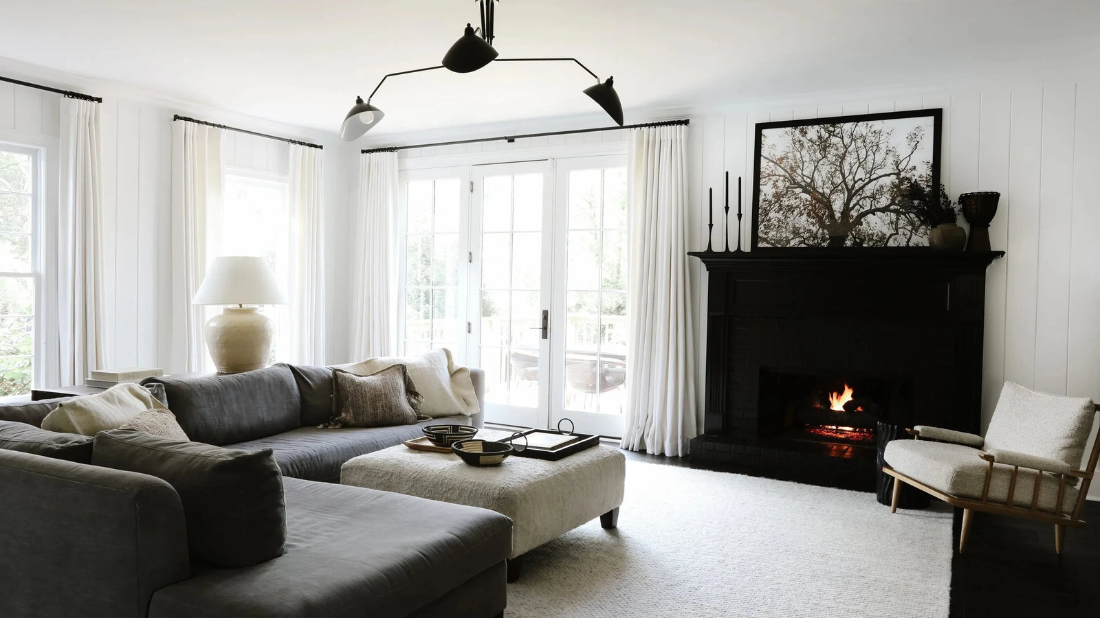 A modern living room featuring a dark gray sectional sofa, a light beige ottoman, a black fireplace with a visible fire, white walls, and large white curtains.