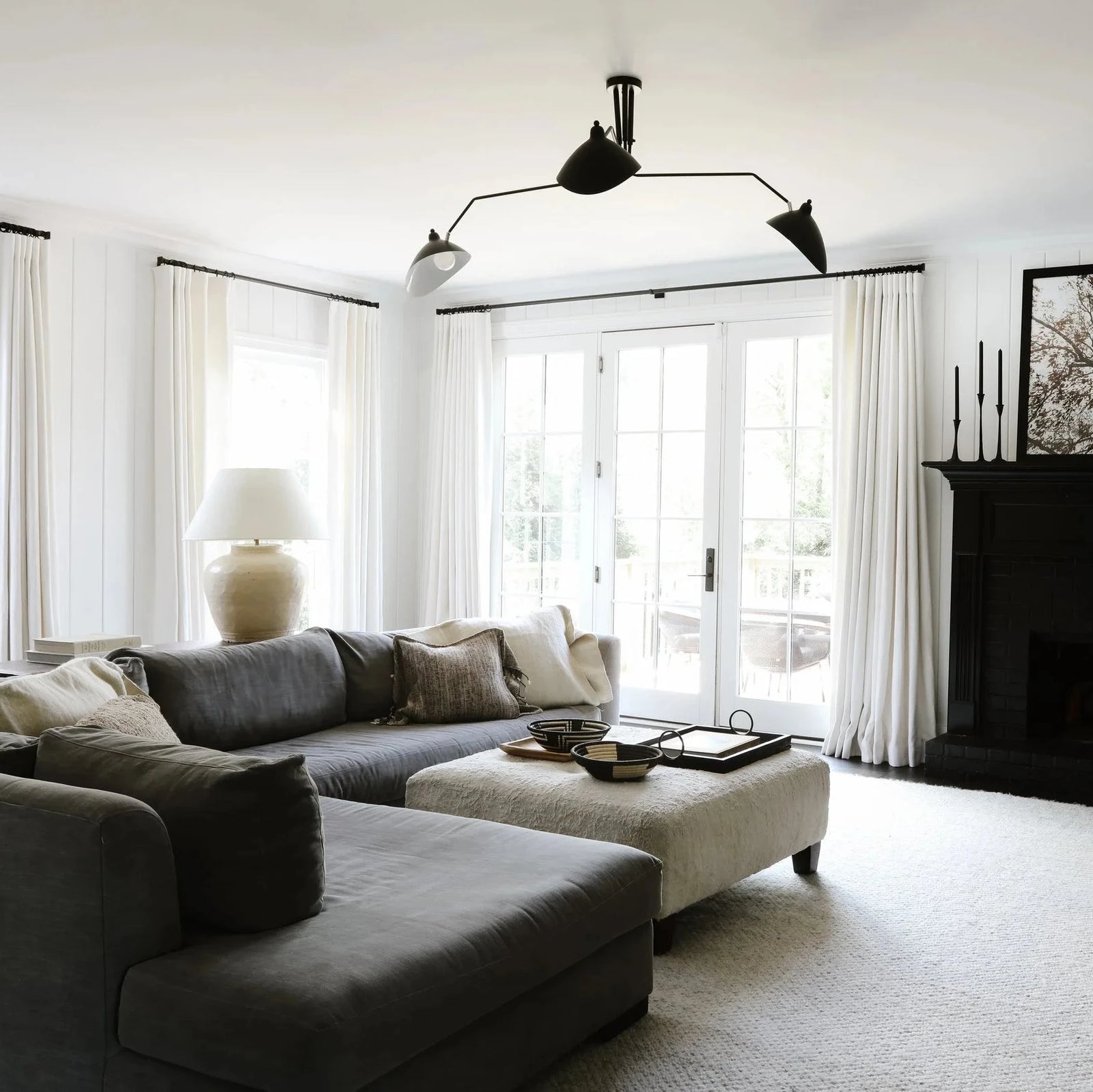 A modern living room featuring a dark gray sectional sofa, a light beige ottoman, a black fireplace with a visible fire, white walls, and large white curtains.